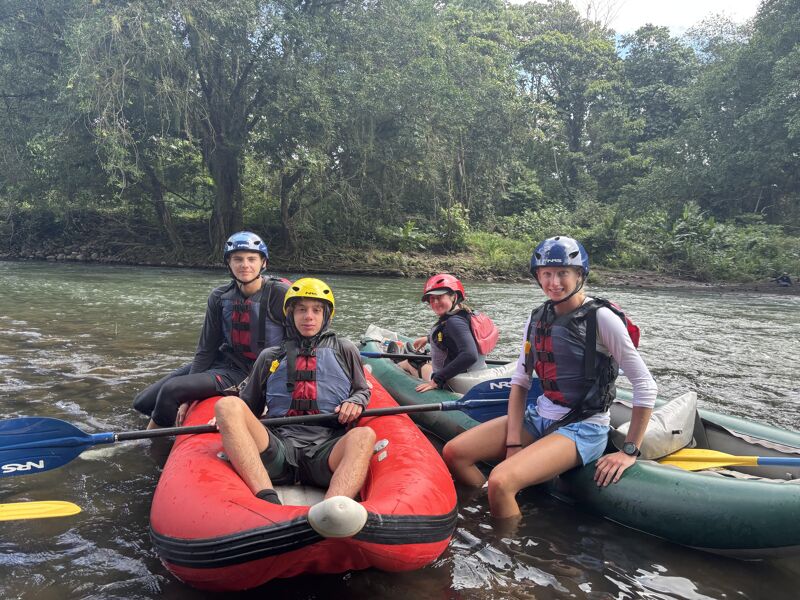 A group of four people are sitting in inflatable rafts on a river, ready for a rafting adventure. They are all wearing helmets and life vests for safety. The rafts are brightly colored, and the river is surrounded by lush green trees. The atmosphere seems cheerful and adventurous, as they prepare to navigate the flowing river.